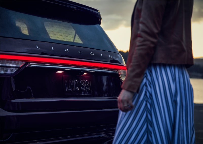 A person is shown near the rear of a 2023 Lincoln Aviator® SUV as the Lincoln Embrace illuminates the rear lights | Lincoln of Wayne in Wayne NJ