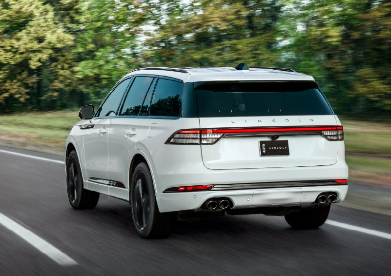 A white 2026 Lincoln Aviator® SUV is being driven away down a road lined with colorful trees. | Lincoln of Wayne in Wayne NJ