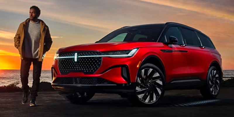 A man wearing a jacket casually walks beside a red Lincoln Aviator SUV against a sunset over the ocean.