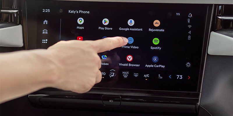 A hand taps Prime Video on a car's infotainment screen displaying various app icons like Maps, Spotify, and Apple CarPlay.
