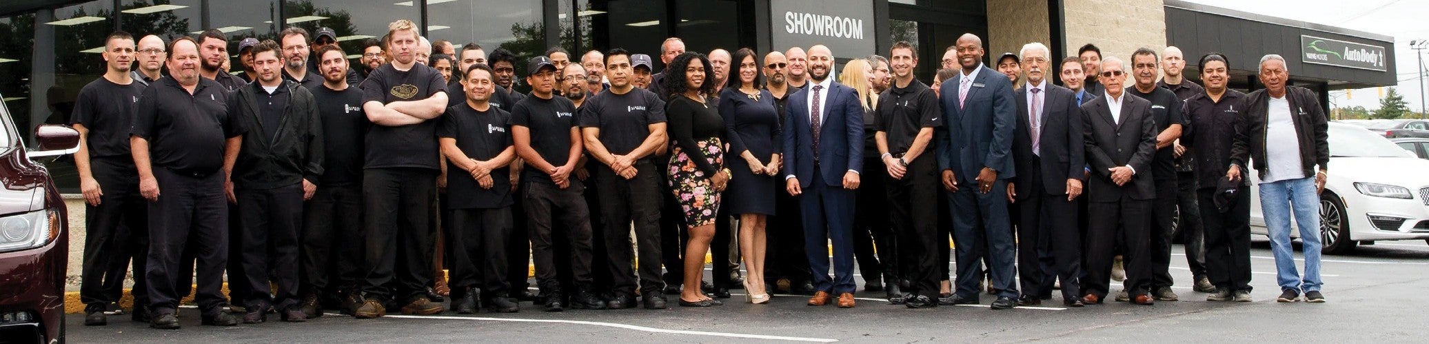 A large group of approximately 30 diverse employees, mostly men, pose for a photo in front of a showroom building.
