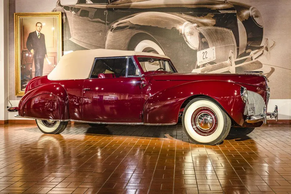 A vintage maroon convertible with a cream soft top, white-walled tires, parked in front of a sepia-toned car mural.
