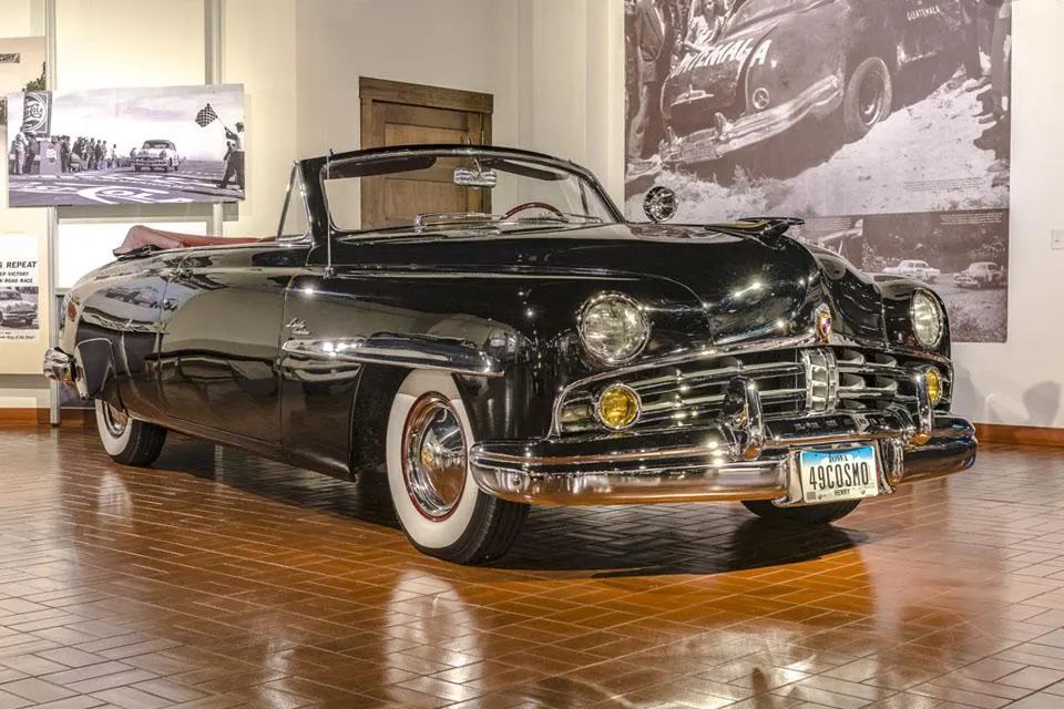 A shiny black vintage convertible with a red interior, white-wall tires, and chrome trim on a tiled floor.