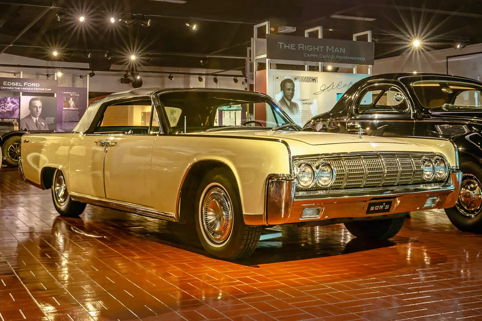A cream-colored 1960s Lincoln Continental convertible with a black top sits on a reflective brick floor.