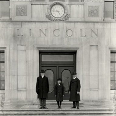 Two men and a small boy in coats and hats stand outside a building with LINCOLN carved above the entrance.