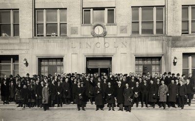 Black and white photo of a very large group of men in coats and hats standing in front of a building entrance.