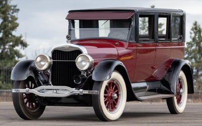 Side view of a vintage dark red car with black trim, fenders, and roof, white-walled tires, and red spoke wheels.