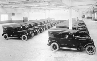 Rows of dark vintage black sedans with canvas tops and running boards inside a large, brightly lit factory.