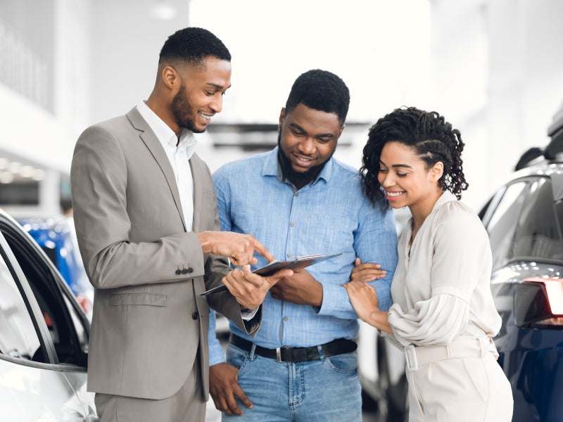 Car salesman showing documents on a clipboard to a smiling couple in a dealership showroom.