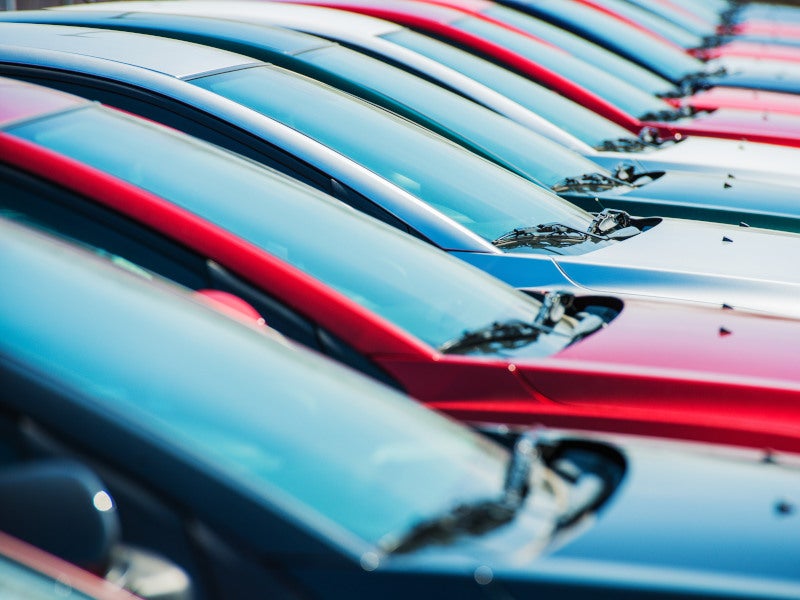 A line of parked cars, alternating red, blue, and silver, showing their hoods and windshields in sunlight.